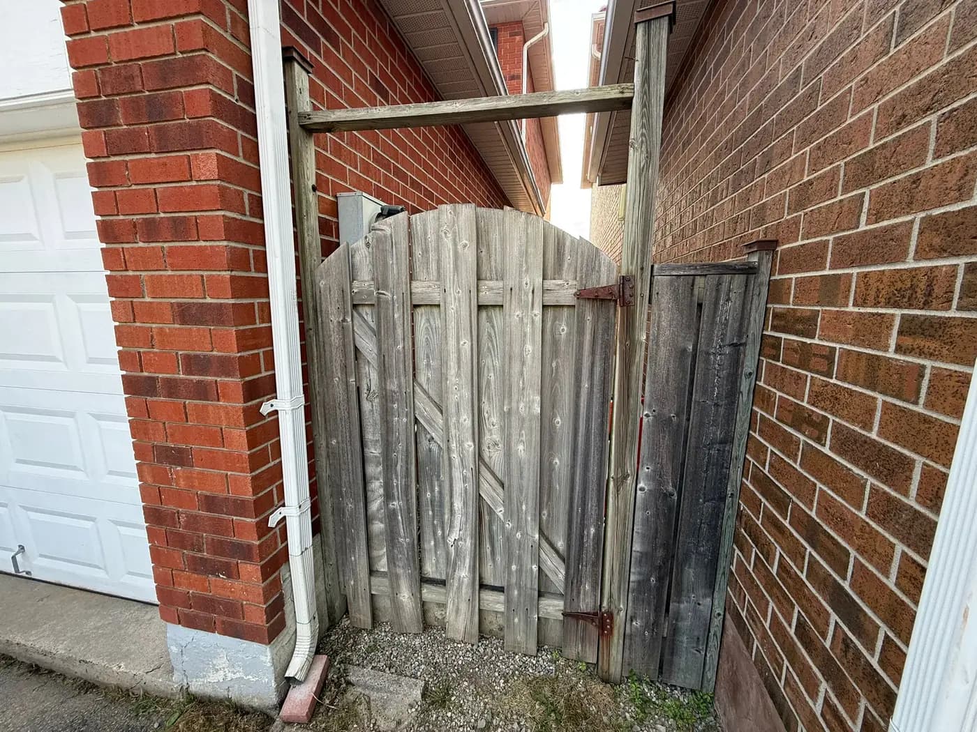 Weathered grey side gate between brick house and garage, warped and hanging off a rotted top frame